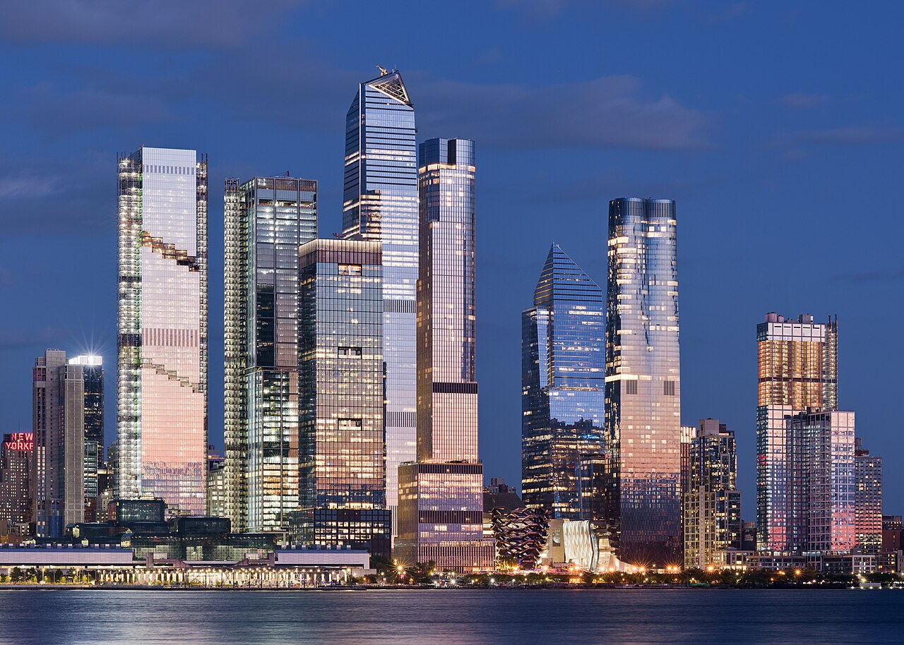 A cluster of glass skyscrapers catching pink evening light above a river, under a darkening blue sky.