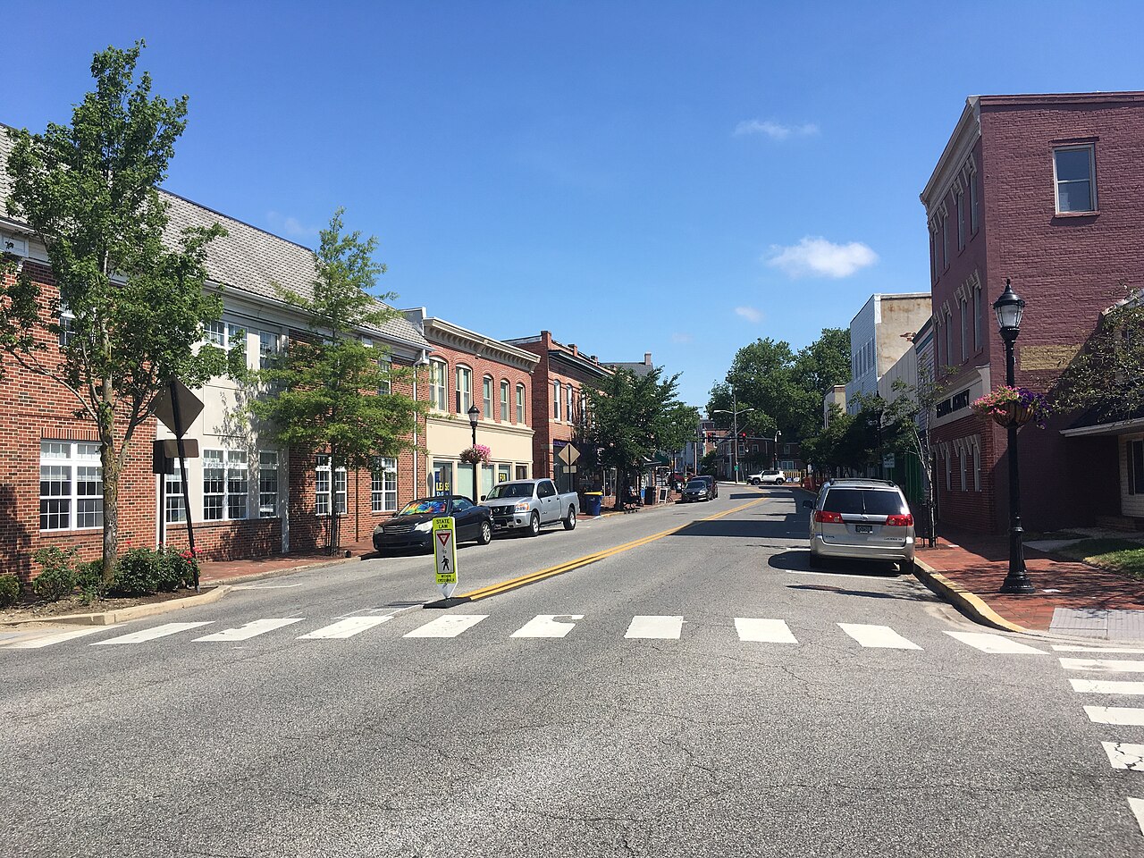 A quiet small-town street with a crosswalk, brick buildings on both sides, street trees and a few parked cars.