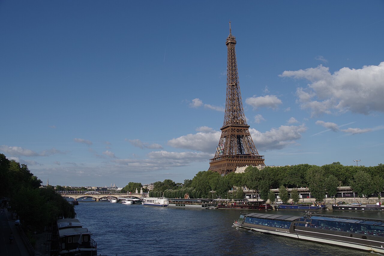 The Eiffel Tower with five coloured rings mounted between its first and second levels, above trees and a river.