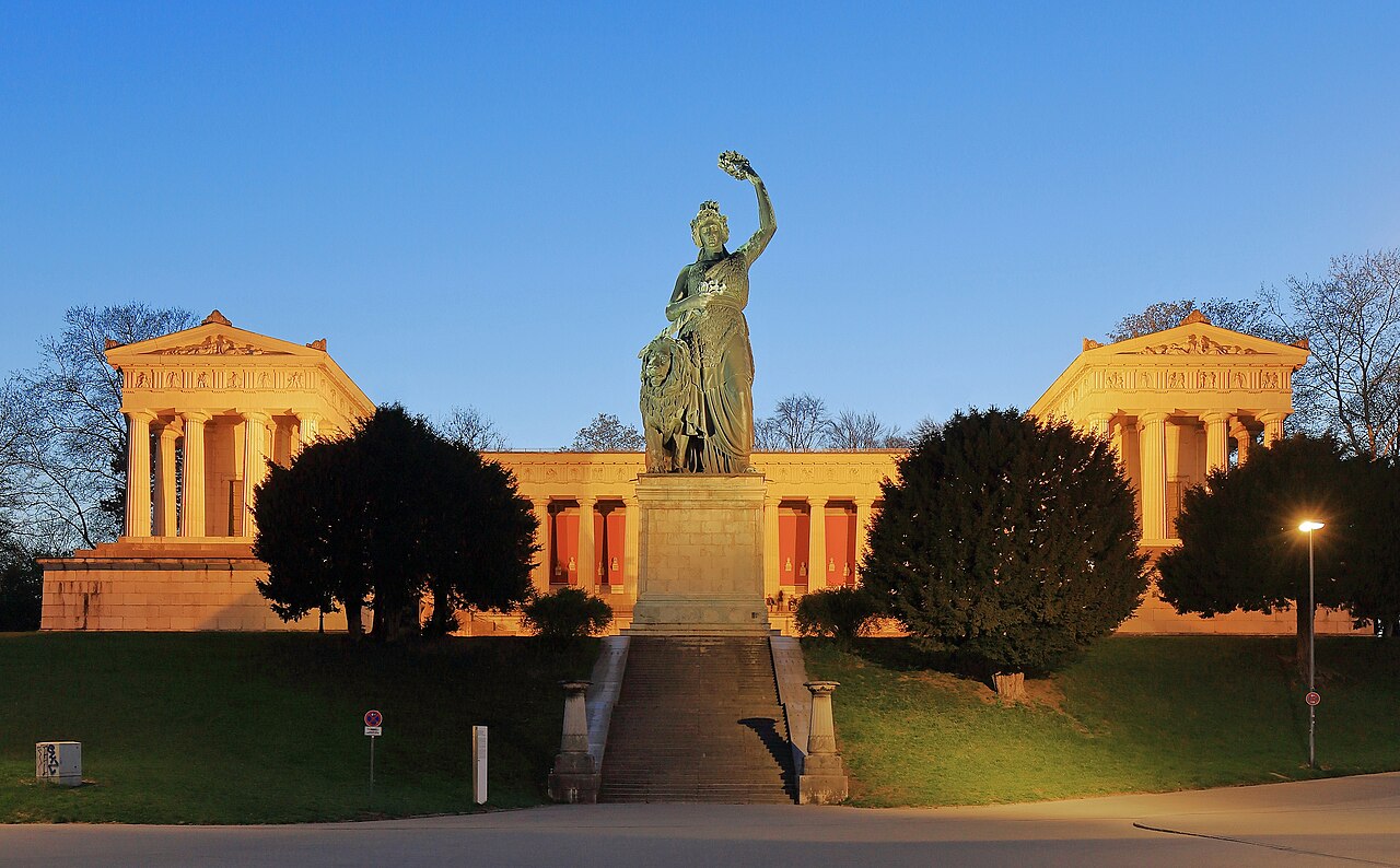 A tall bronze statue of a woman with a raised arm in front of a columned hall, lit warm by low sun between dark trees.