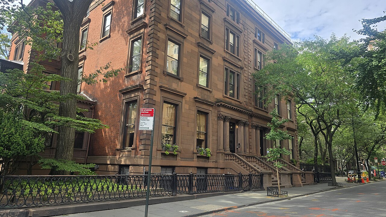 A four-storey brownstone house on a corner of a tree-lined street, with an iron fence along the pavement.