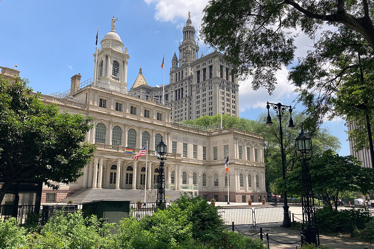A low pale stone city hall with a cupola, a much taller ornate tower behind it, framed by green trees.