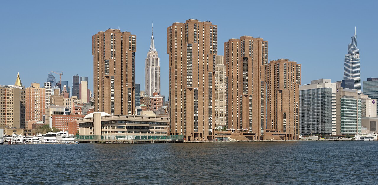 Four brown brick apartment towers standing at the edge of a river, with a tall spired skyscraper between them.