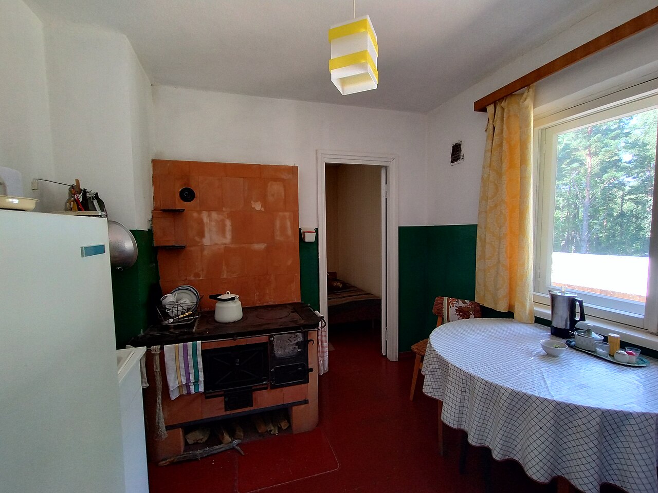 A small kitchen with a rounded pale fridge, an orange tiled stove, a round table with a checked cloth and a window with yellow curtains.
