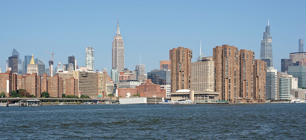 Brown brick apartment towers stand at the edge of a wide river with a tall spired skyscraper rising behind them.