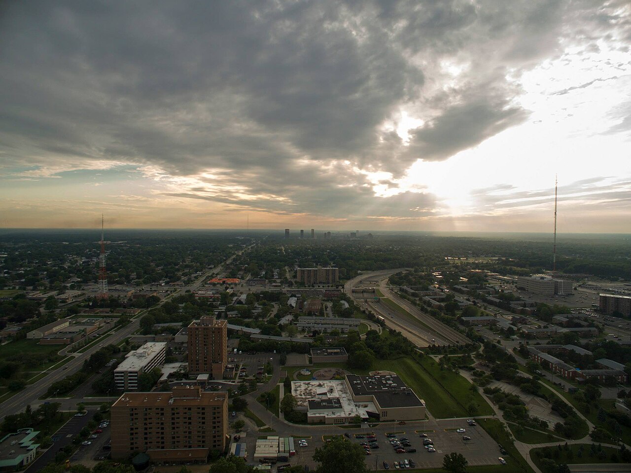 An aerial view of a flat, tree-covered suburb crossed by a motorway, under heavy clouds with the sun breaking through.