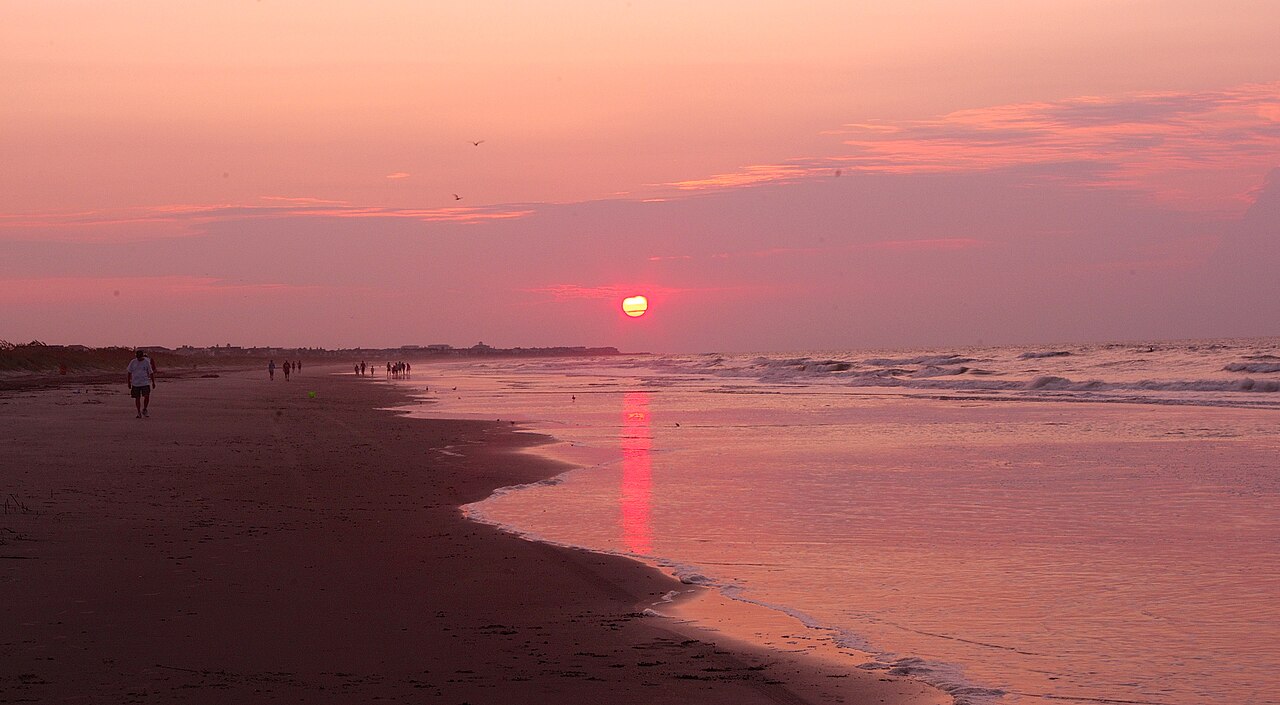A red sun sits low over the sea, colouring the sky and the wet sand of a long beach pink.