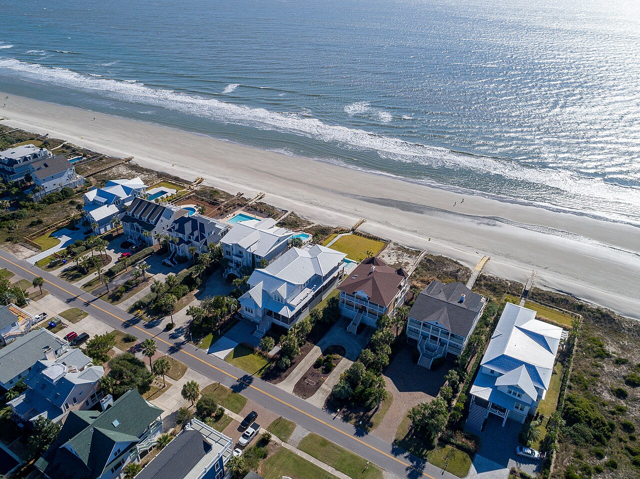 Rows of large beach houses with pale roofs line a road beside a wide sandy beach and breaking surf.