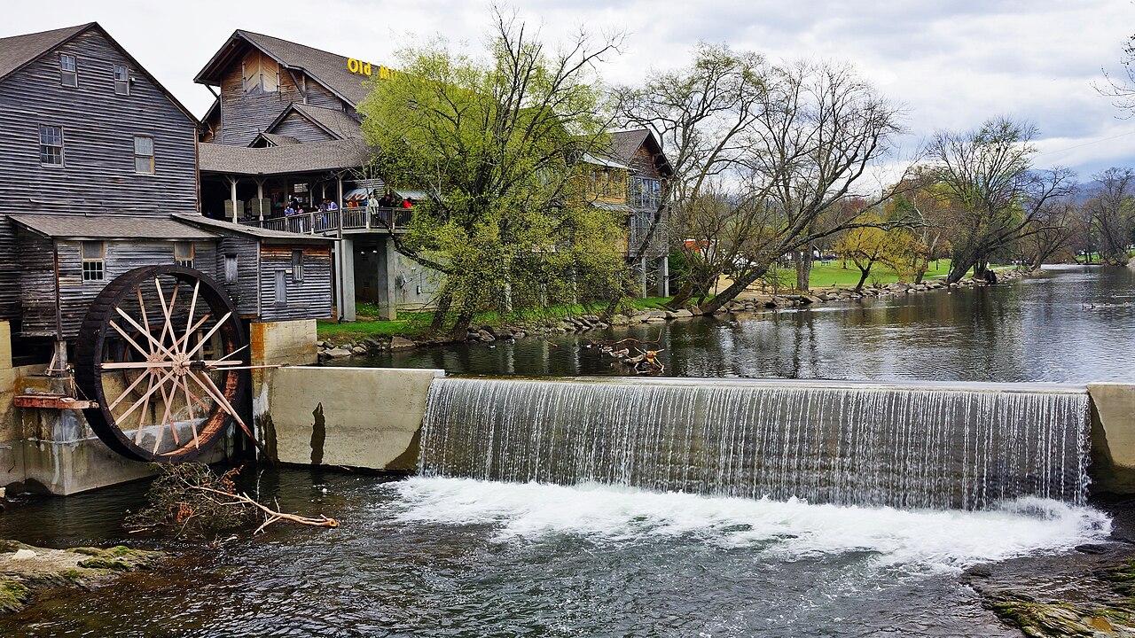 A weathered wooden mill with a large water wheel stands beside a low dam where the river spills over in a white sheet.