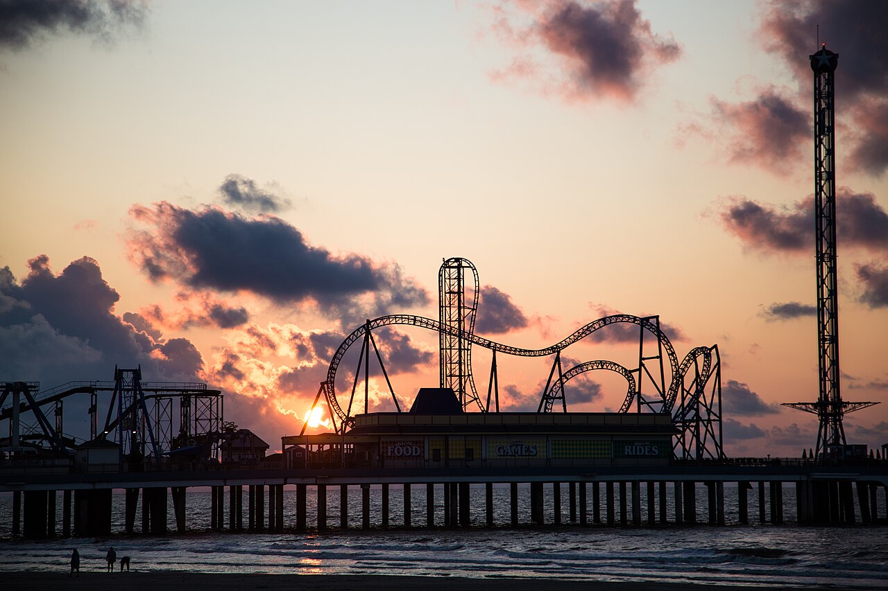 The dark outline of a pier carrying a roller coaster and a tall tower ride against an orange and purple evening sky.