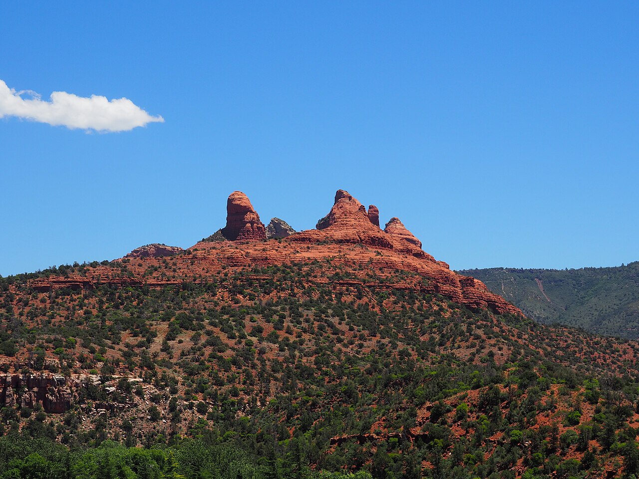 A jagged red sandstone butte rises from a slope dotted with green shrubs under a clear blue sky.