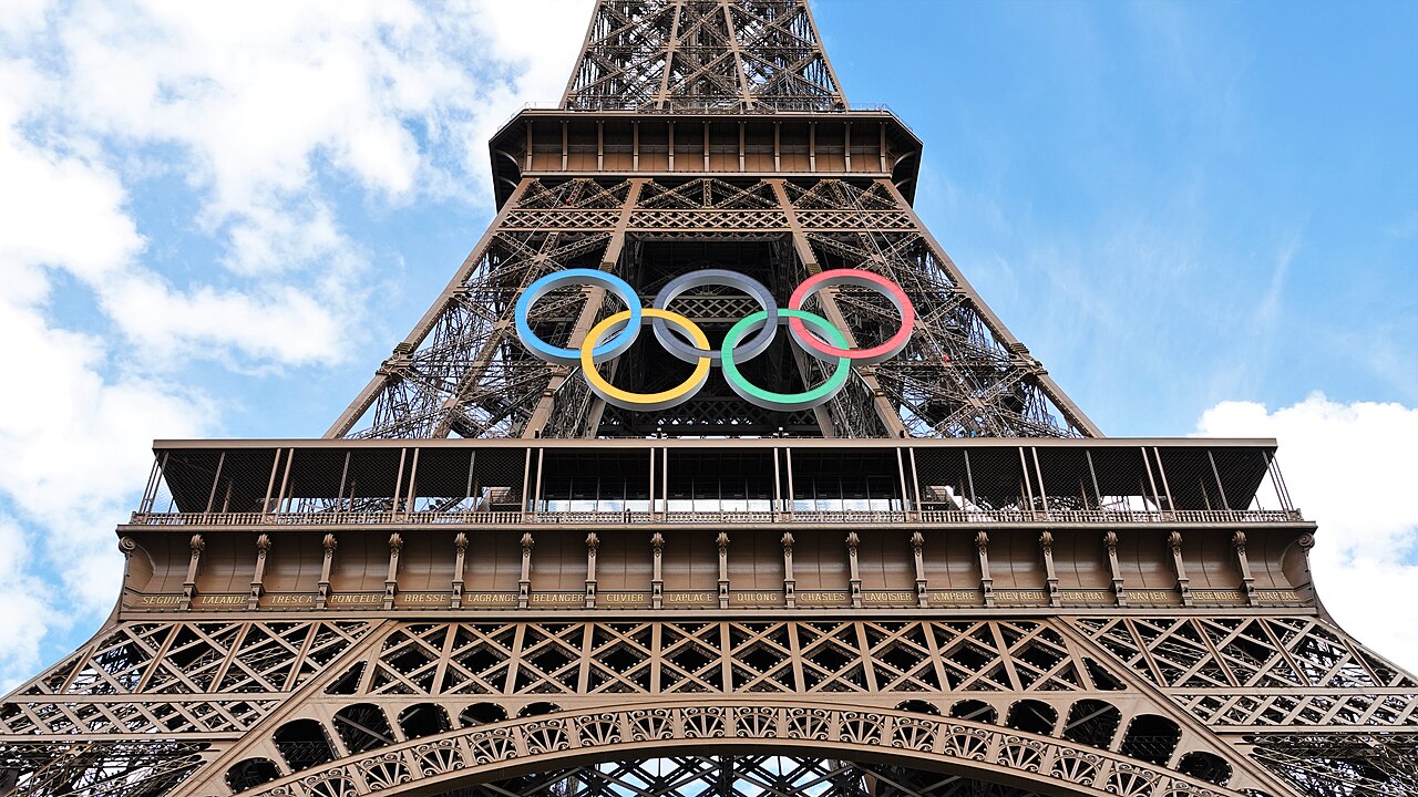 Five coloured rings hang on the iron lattice of a tower, seen from directly below against a blue sky.