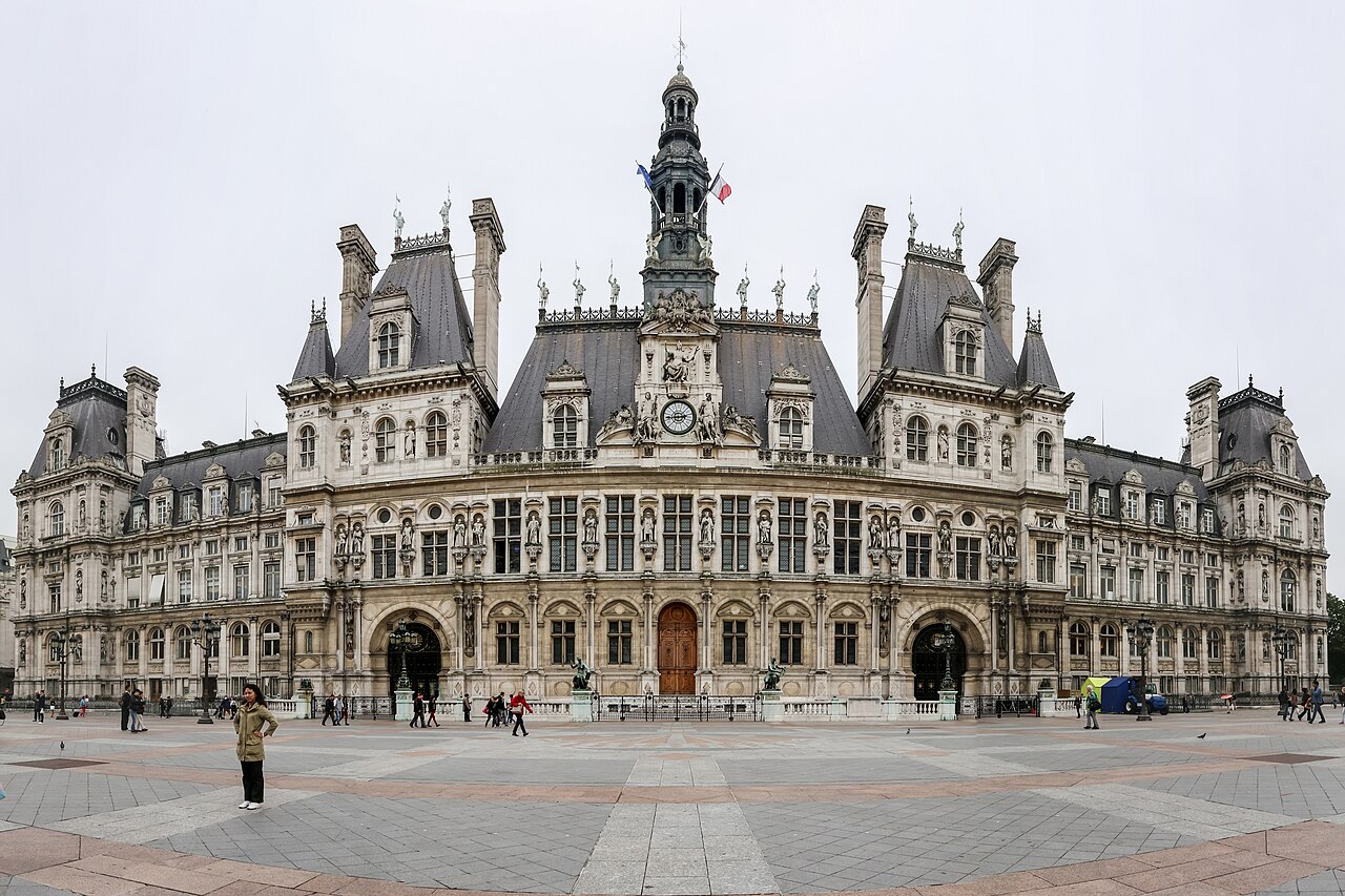 A wide, ornate stone building with steep slate roofs and a central clock tower stands behind an open paved square.
