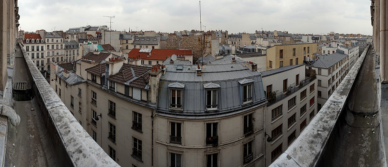 Grey zinc mansard roofs, dormer windows and chimney pots top a block of pale stone Paris apartment buildings.