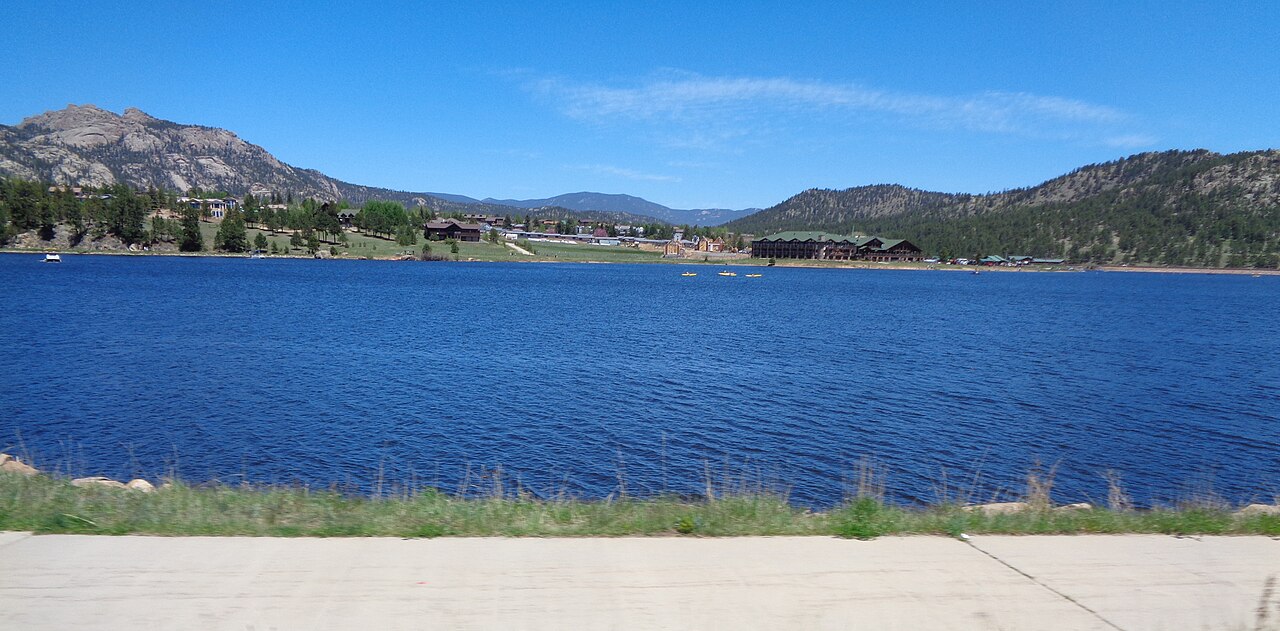 A deep blue lake lies below low rocky hills, with buildings along the far shore and a clear blue sky.