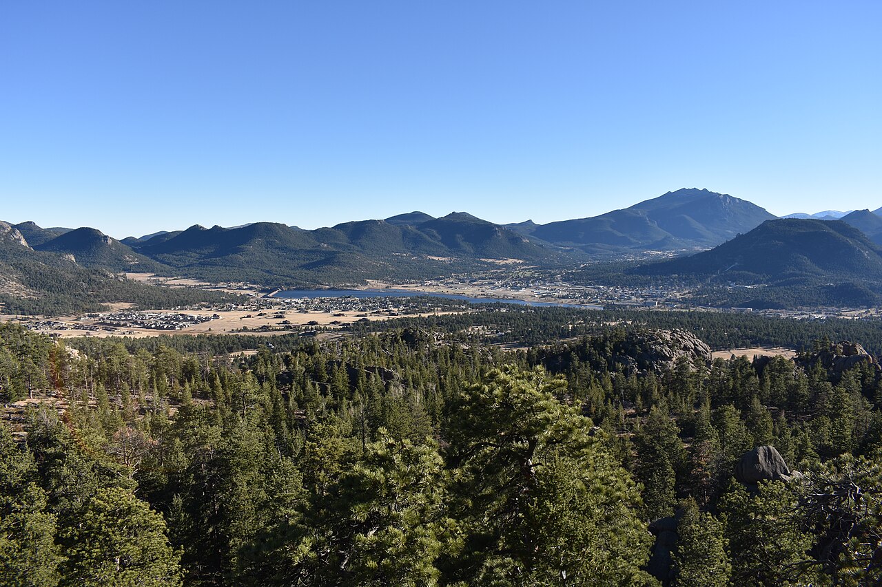 A town spreads across a valley floor below pine-covered slopes, with a range of blue mountains behind.