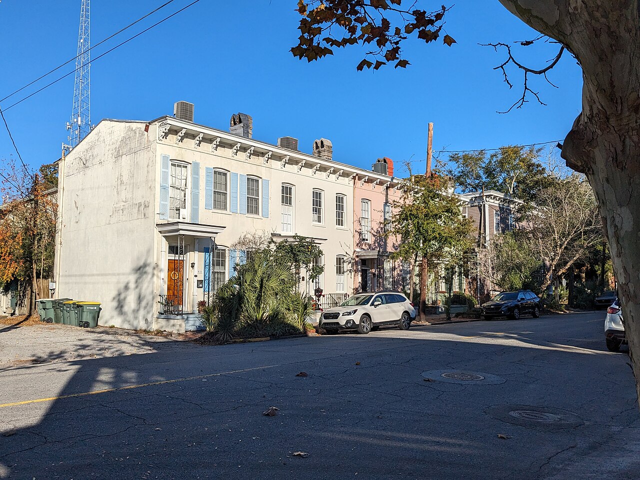 A terrace of pastel-painted two-storey houses with shutters stands on a street corner in bright sun, with cars parked along the kerb.