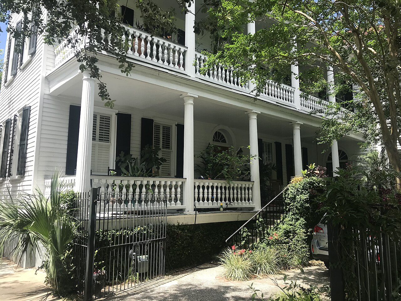 A white wooden house with two storeys of columned side porches and balustrades, behind an iron fence and palms.