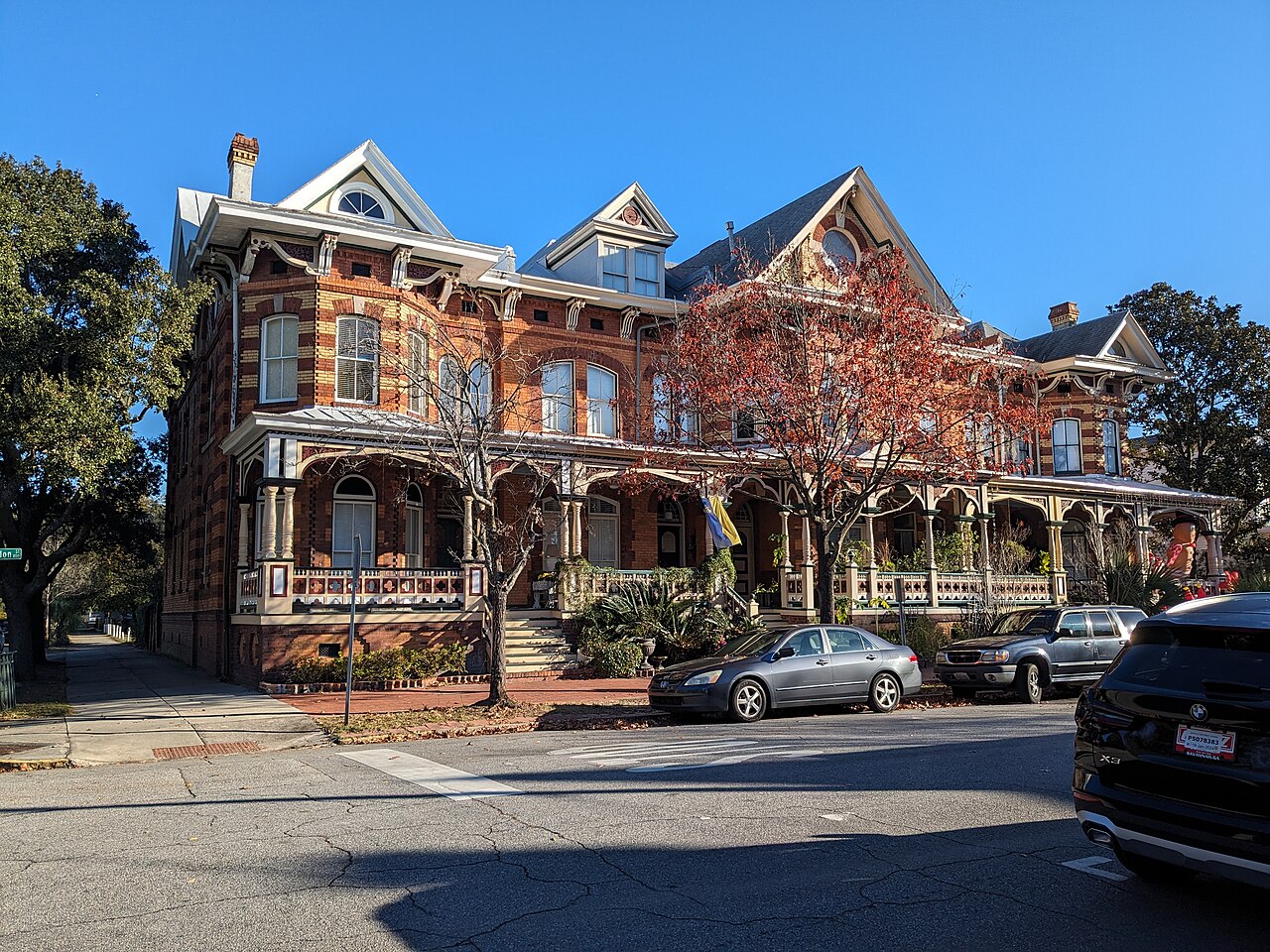 A long red-brick Victorian terrace with gables and a wooden porch along its front, with cars parked on the street beside it.