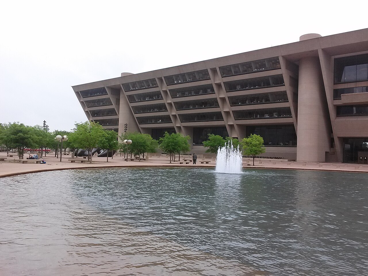 A wide concrete building that leans outward as it rises stands behind a pool with a fountain and a row of trees.