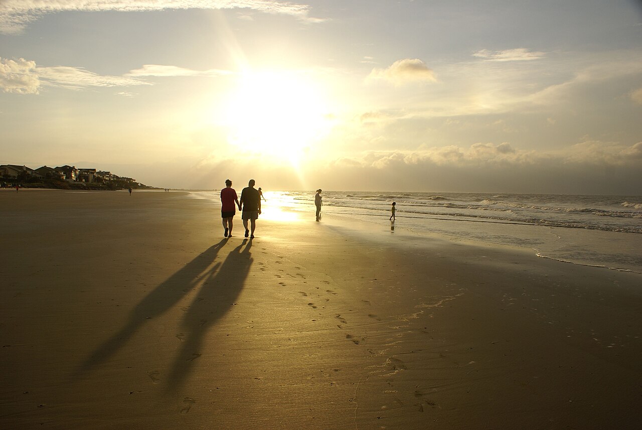 A broad sandy beach with the sun low behind haze, long shadows on the sand and a few distant walkers in silhouette.