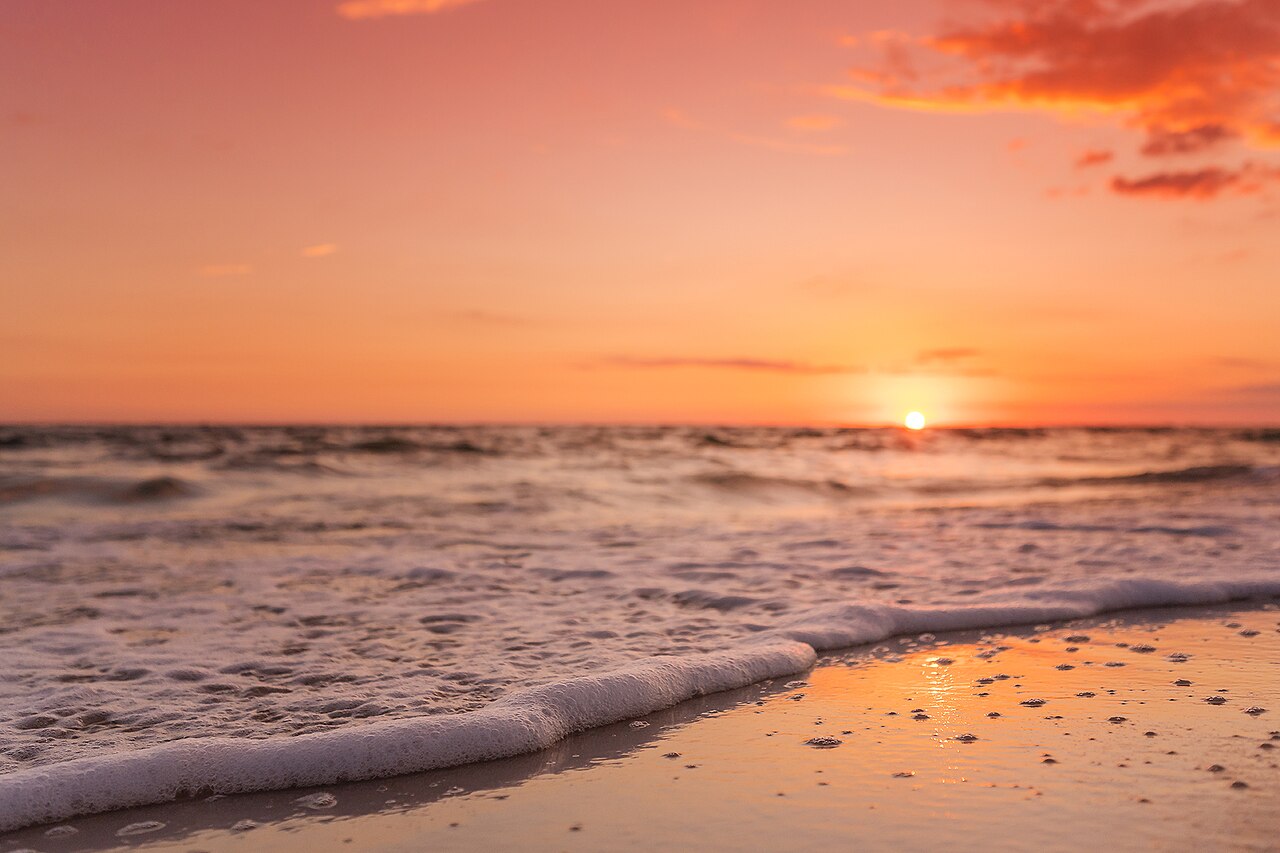 Small waves washing onto wet sand under an orange and pink sky, with the sun touching the horizon.