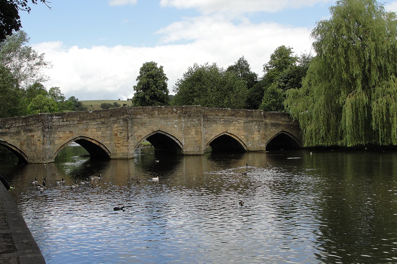 A low stone bridge with five pointed arches crossing a calm river, with ducks on the water and willows on the far bank.