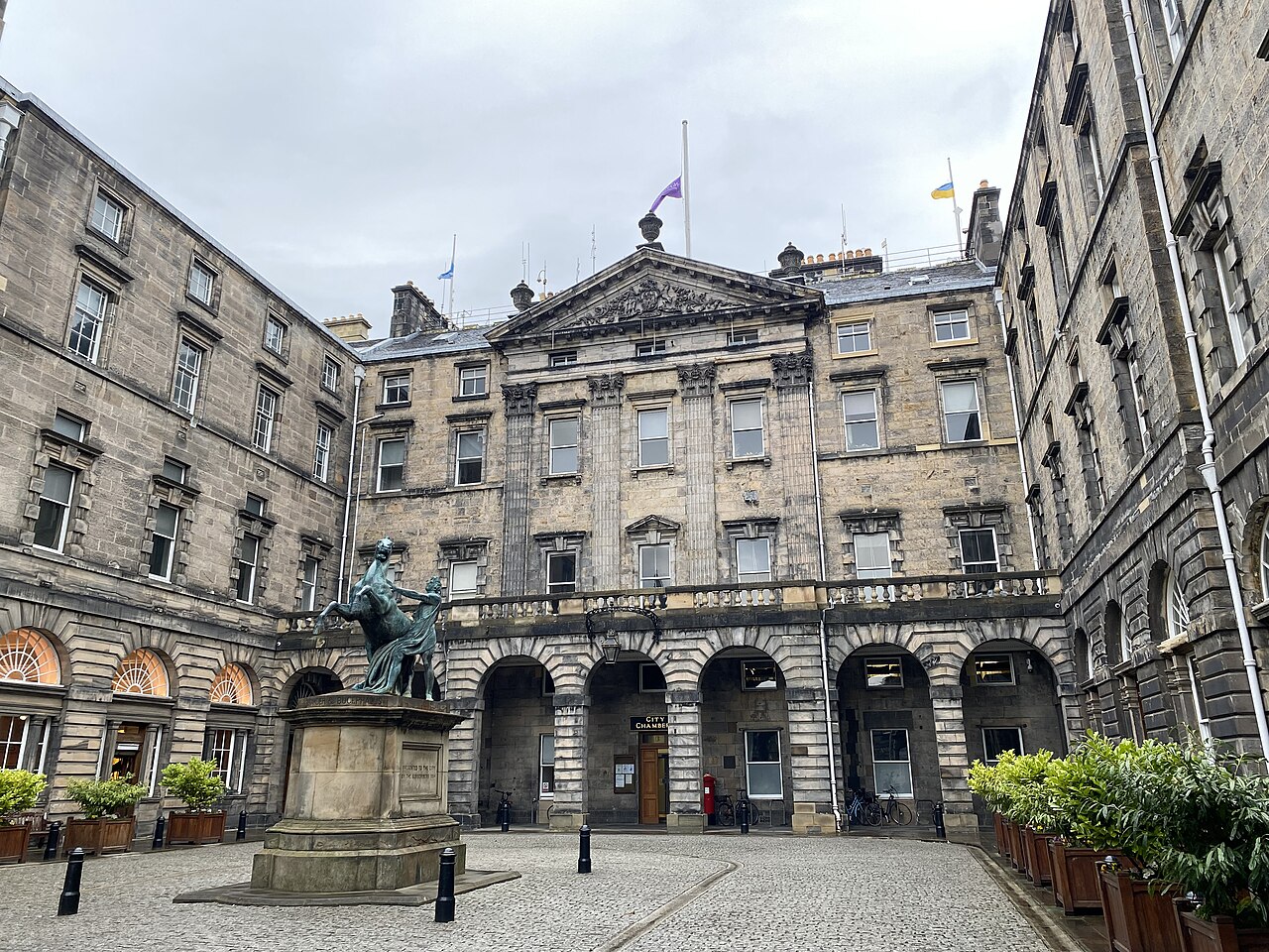 A grey stone courtyard with arched arcades on three sides, a bronze statue on a plinth in the centre and a flag on the roof.