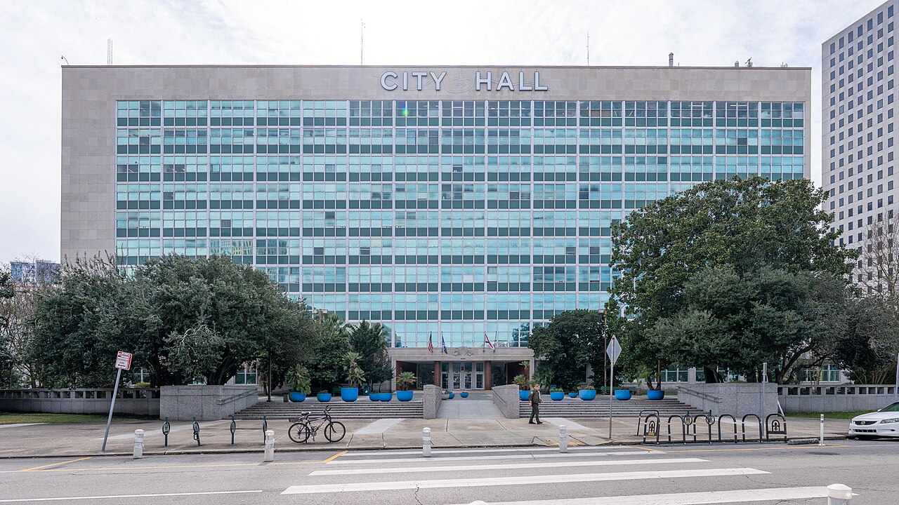 A wide mid-century office block with a blue-green glass front and the words City Hall along the top, behind trees and a paved plaza.