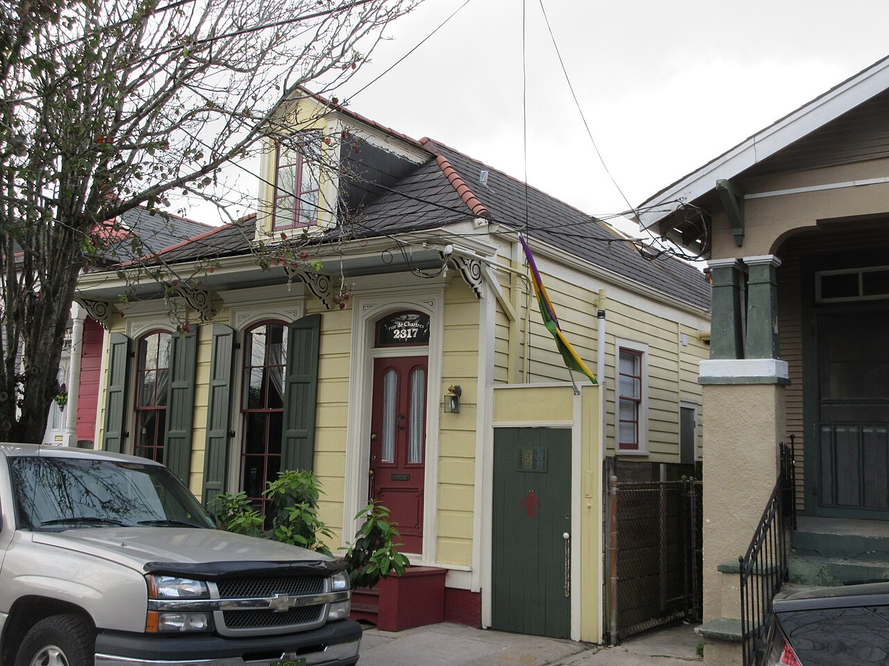 A pale yellow wooden cottage with green shutters, a red front door and a dormer window, beside a neighbouring house with a porch.