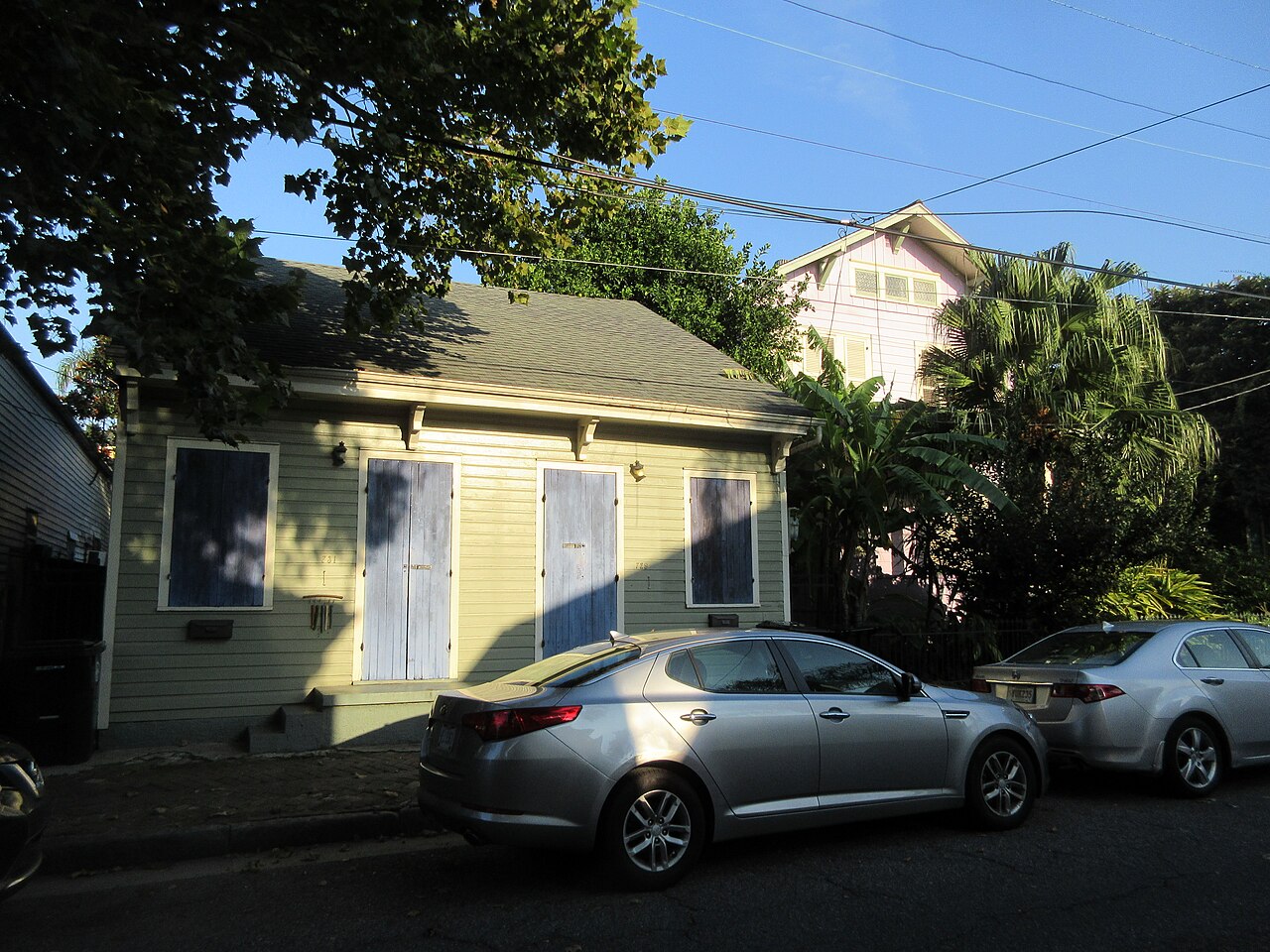 A small yellow wooden cottage with shuttered windows and a pitched roof on a tree-shaded street, with cars parked at the kerb.