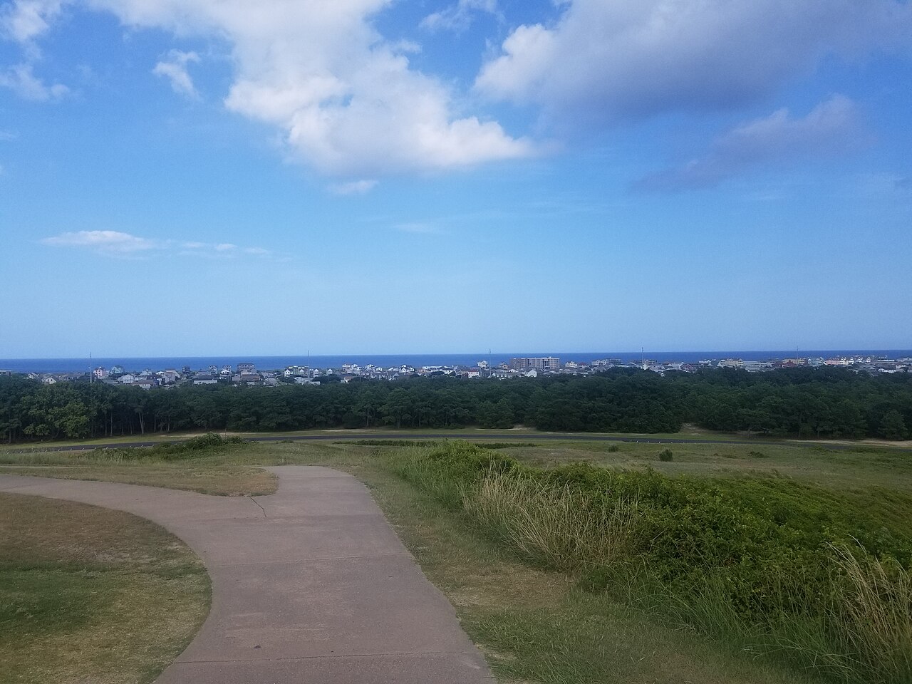 A path curving down a grassy hill towards low woods, a line of buildings and the sea in the distance.