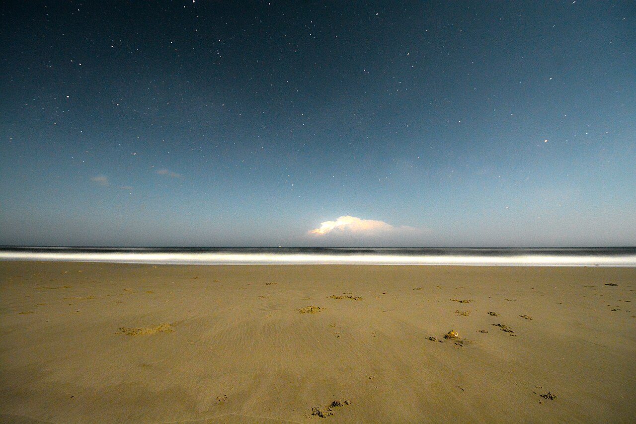 A wide empty sand beach under a starry dark blue sky, with one bright storm cloud low on the sea horizon.