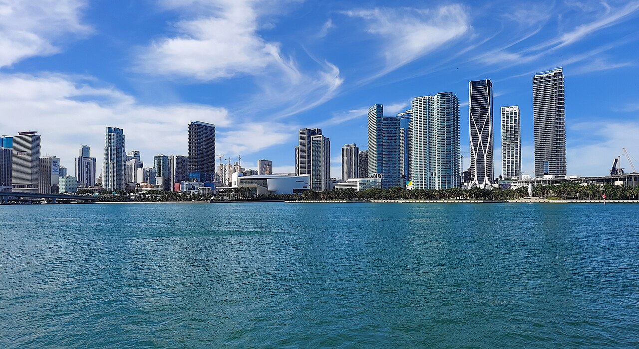 A row of glass towers along the shore behind choppy turquoise water, under a blue sky with streaked white cloud.