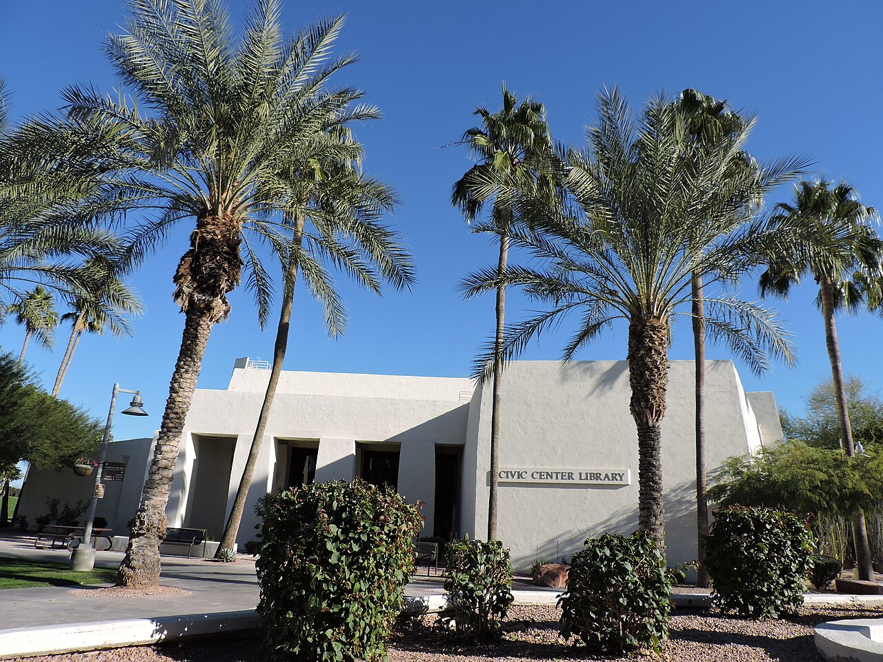 A low white modern building behind tall palm trees and clipped shrubs under a deep blue sky.