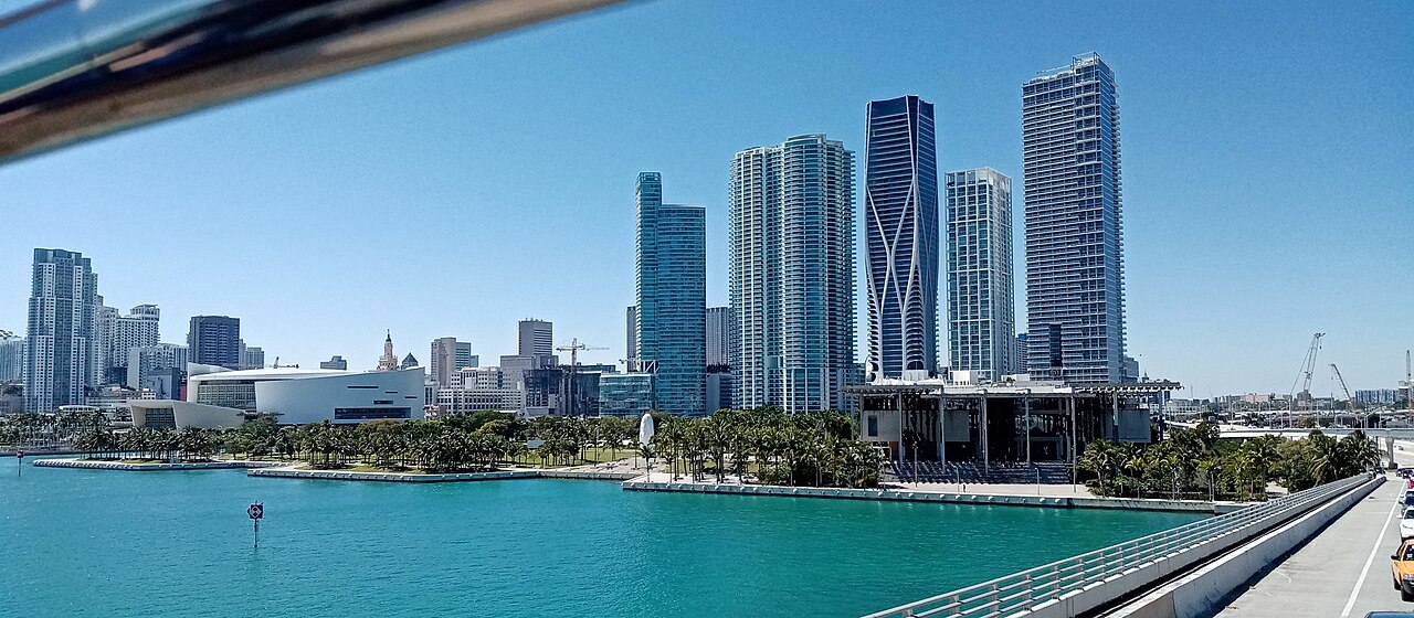 Tall residential towers behind a waterfront park and turquoise water on a clear day.