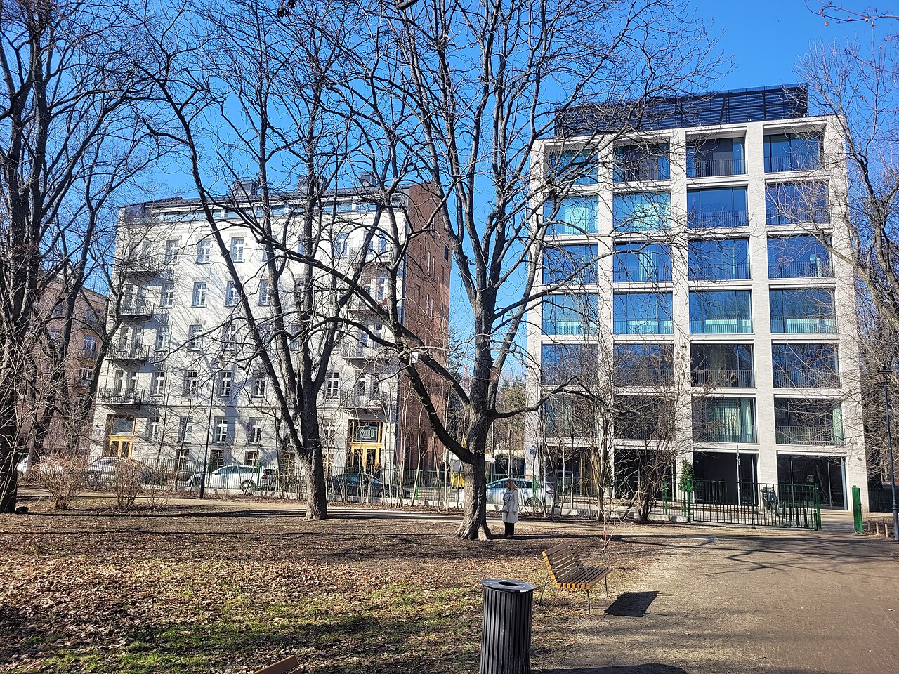 A modern apartment block with a white grid facade beside an older pale building, seen through bare winter trees.