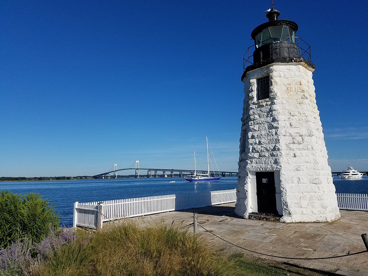A white stone lighthouse with a black lantern at the water's edge, a long suspension bridge crossing the bay in the distance.