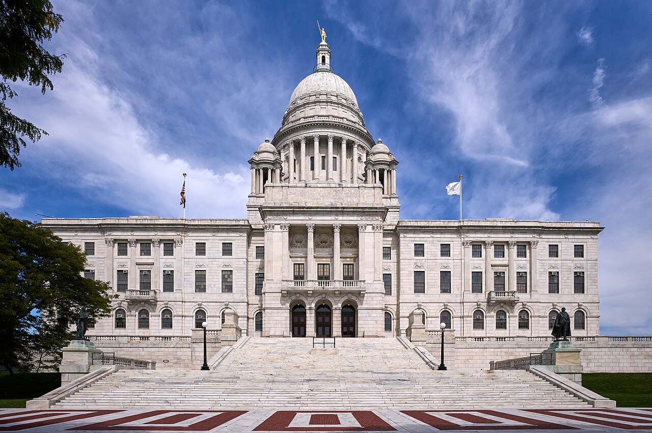 A white marble government building with a central dome and columned entrance, seen across a paved forecourt under a blue sky with thin cloud.