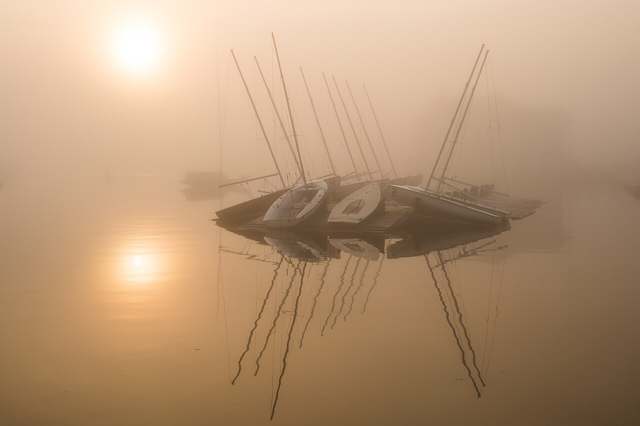 Small sailboats resting on a dock in still water, half hidden in orange fog with a pale sun above and its reflection below.