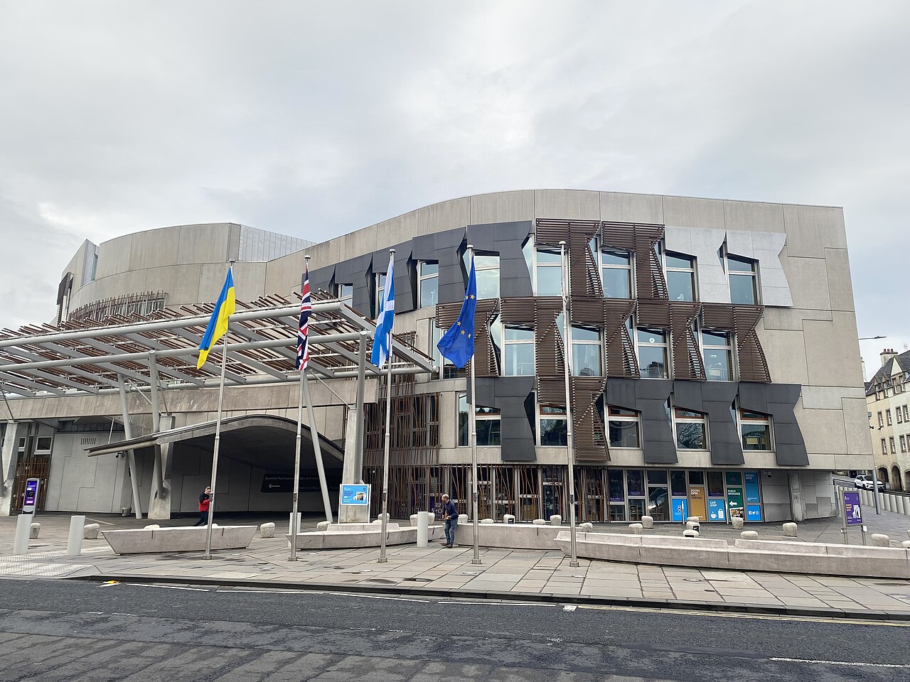 A modern building with dark angular window panels and a curved roof behind a row of flagpoles, across a wide paved square.