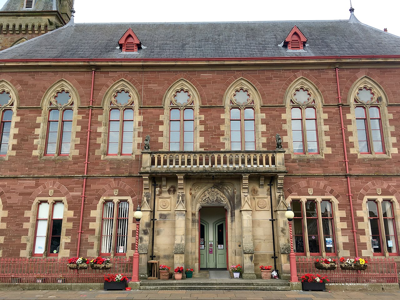 The front of a red sandstone hall with a row of arched upper windows, a stone balcony over the door and flower boxes along the ground floor.