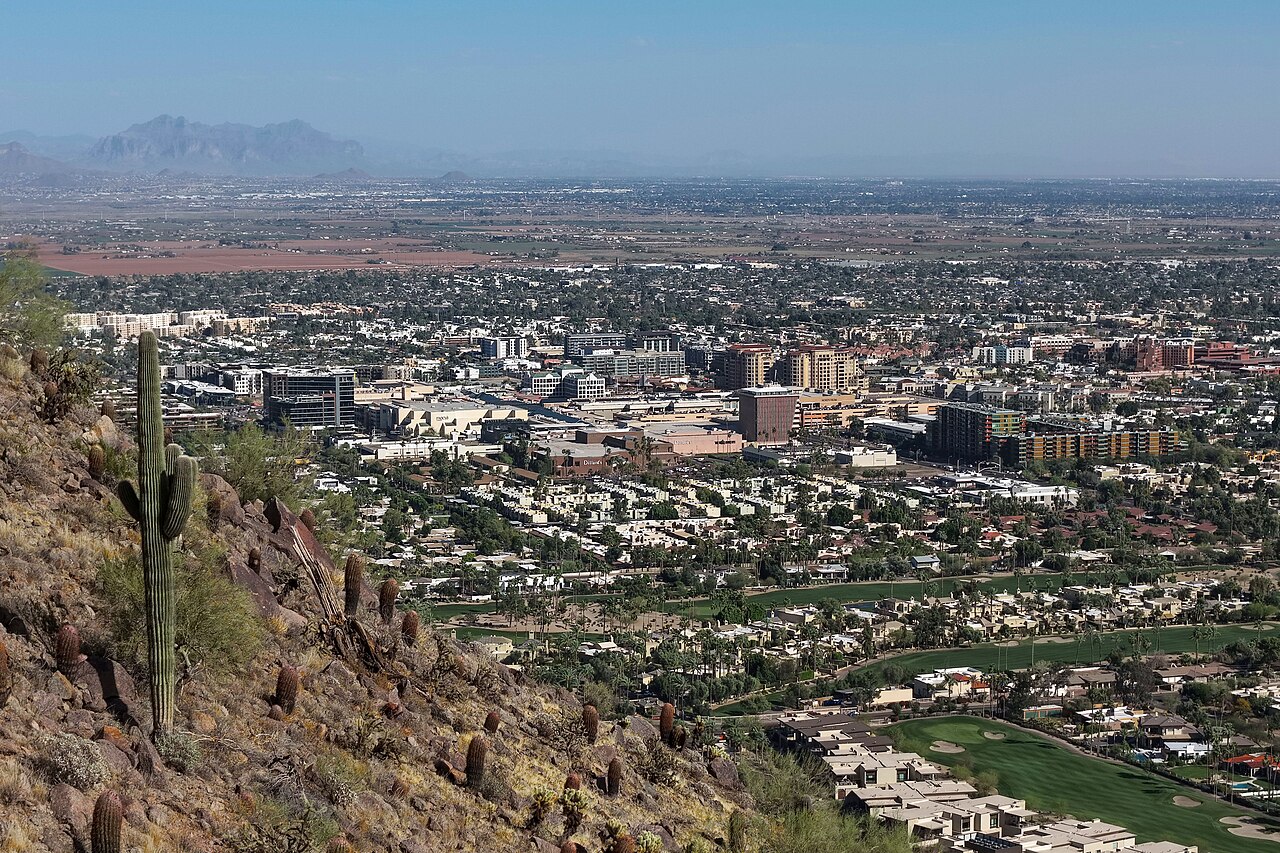 A wide view from a rocky desert slope with a saguaro cactus down onto a flat city of low buildings and a few mid-rise blocks, hazy mountains beyond.