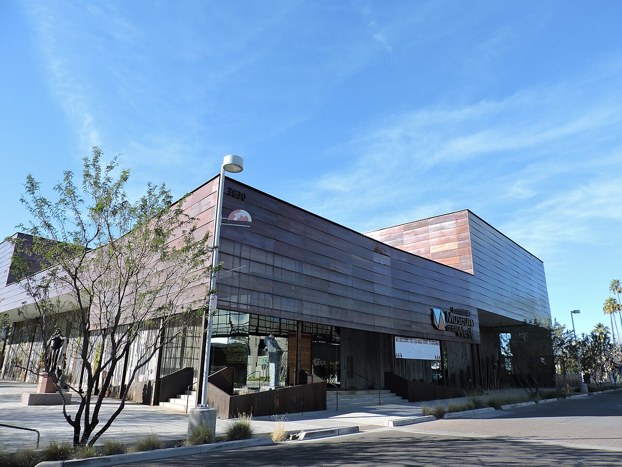 A modern two-storey building clad in dark ribbed metal on a street corner, with a bare desert tree in front and a deep blue sky.