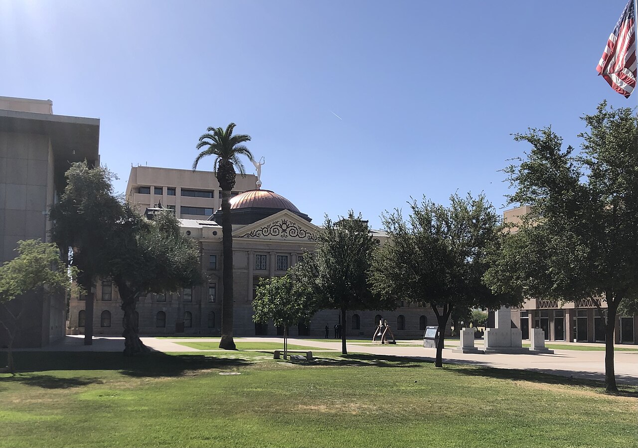 A low domed building seen across a shaded lawn with palm and leafy trees, a flag hanging at the top right.