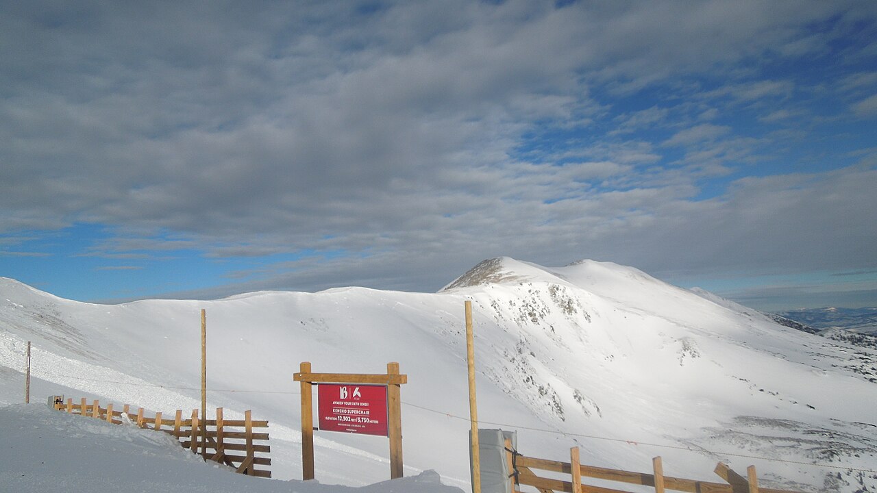 Bare snow-covered peaks under a cloudy sky, with a wooden fence and a red trail sign in the snow in the foreground.
