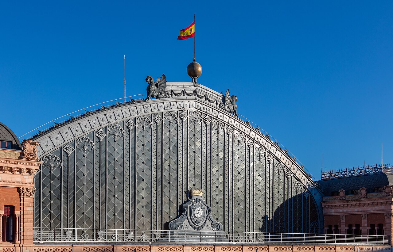 A large arched iron and glass station roof between red brick wings, a Spanish flag flying above it against a clear blue sky.