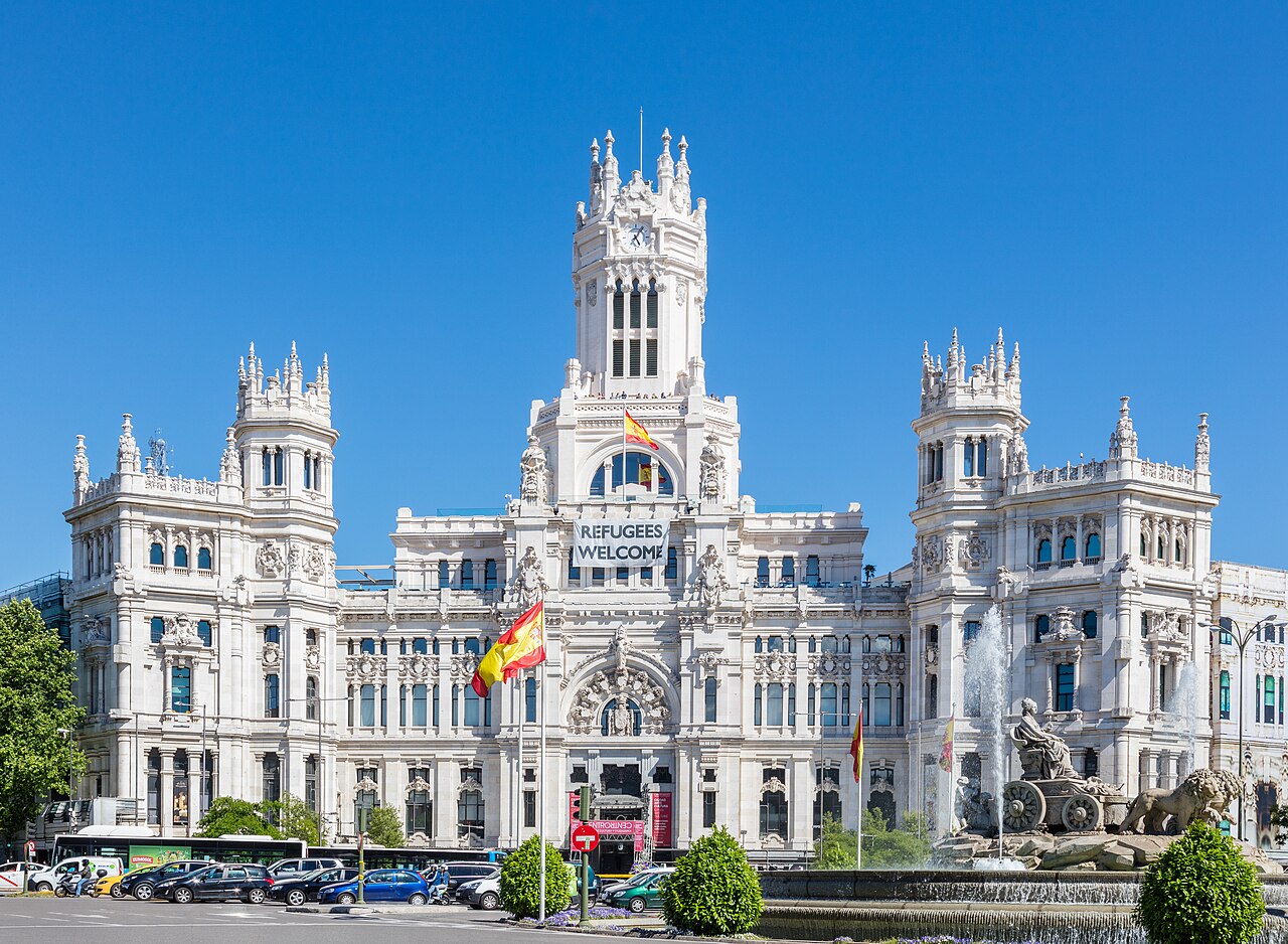 A wide white stone palace with a tall central clock tower and pinnacled side towers under a deep blue sky, Spanish flags on its front.