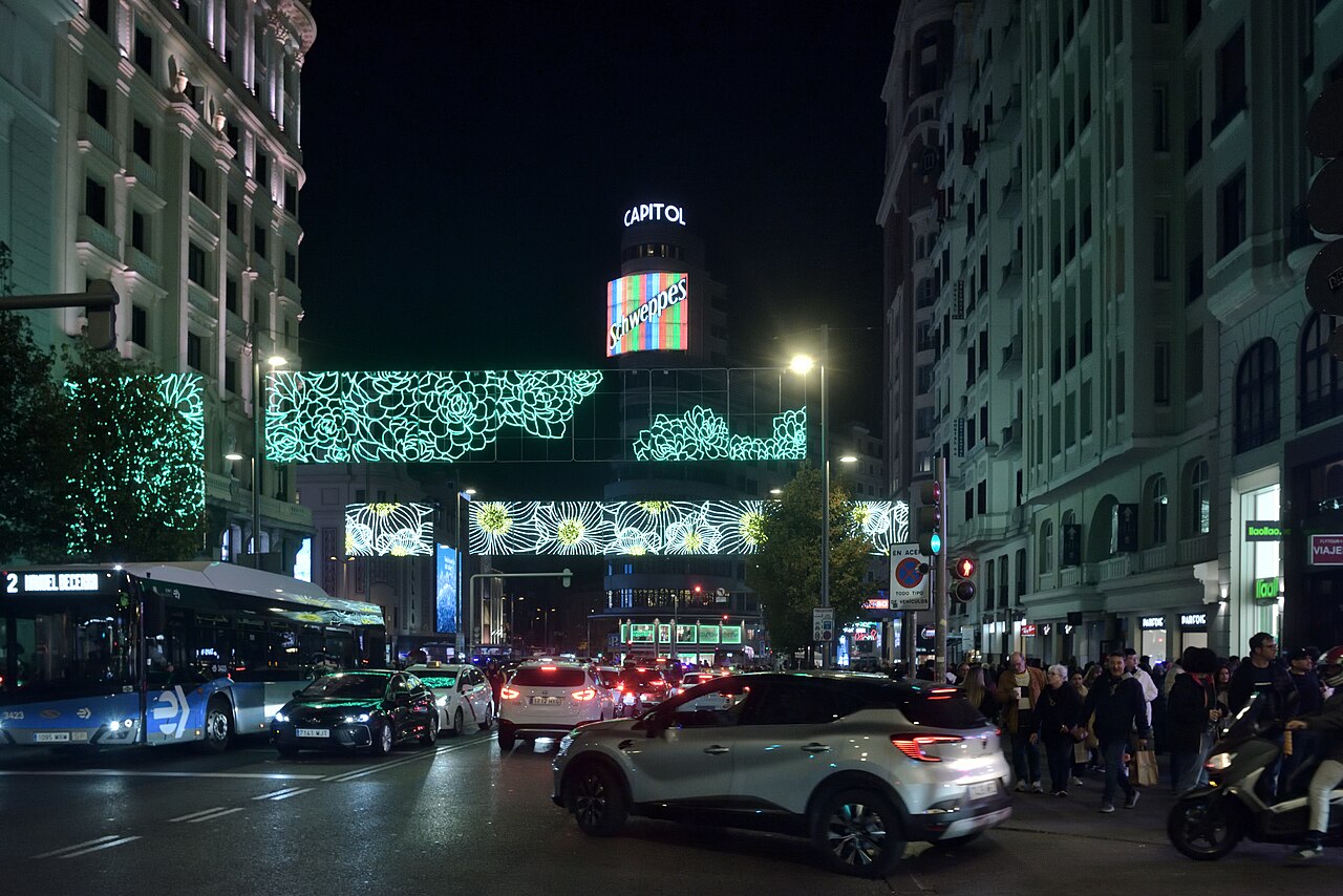 A busy city avenue at night with cars and buses, tall buildings on both sides and strings of green lights hung across the street.