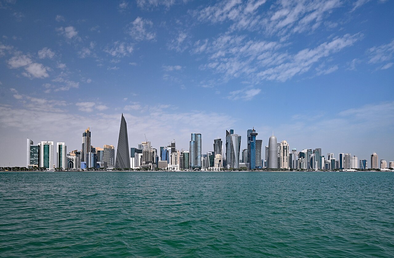 A long row of modern towers along a shoreline, seen over green sea water under a blue sky with thin clouds.